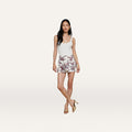 A woman stands against a plain white background, posing with one leg crossed over the other and her hands relaxed by her sides. She is wearing a white tank top, tan heeled sandals, and the Cyra Low-Rise Mini Skirt in a floral pattern.