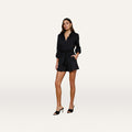 A woman stands against a plain light background, wearing the Damira Poplin Short as part of a black long-sleeve belted romper made from stretch poplin with ultra-high waist shorts and black heeled sandals, her hand casually placed in her pocket.