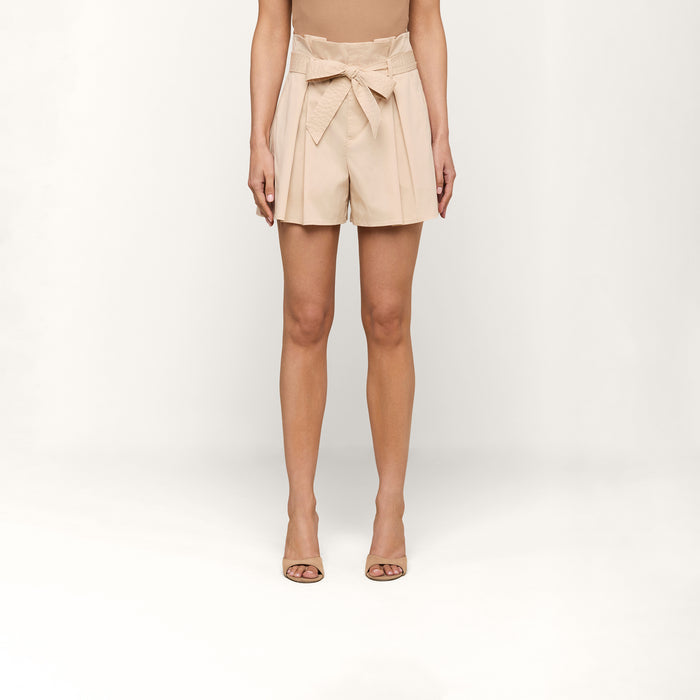 A woman stands against a plain white background wearing the Damira Poplin Short—beige ultra-high waist shorts featuring a self-tie belt and pleats—paired with a tan top and open-toed heel sandals.
