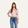 A woman with long brown hair is seen standing against a plain white background, wearing the Dani Silk Blouse in light pink—crafted from 100 silk charmeuse—paired with blue jeans and a brown belt.