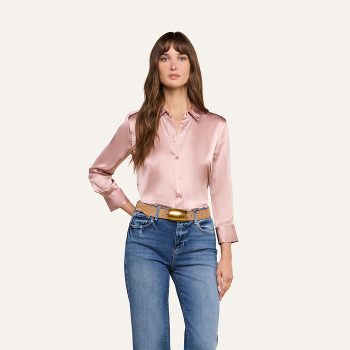 A woman with long brown hair is seen standing against a plain white background, wearing the Dani Silk Blouse in light pink—crafted from 100 silk charmeuse—paired with blue jeans and a brown belt.