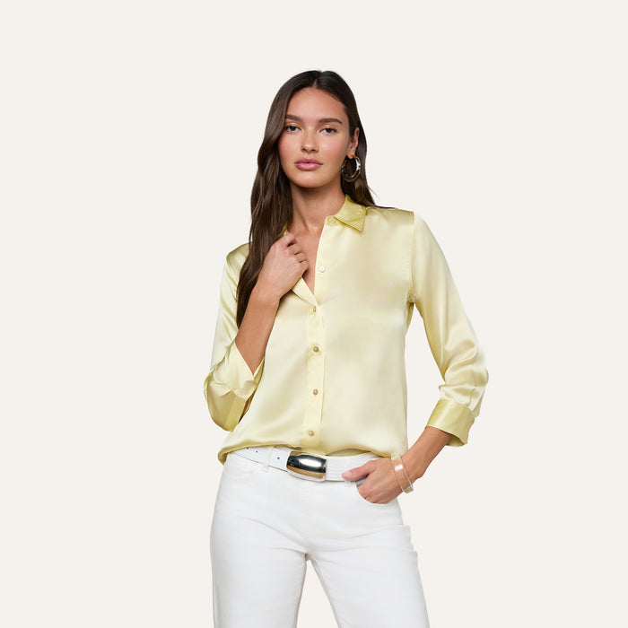 A woman wearing the Dani Silk Blouse in light yellow and white pants stands against a plain light background, with one hand touching her collar and the other in her pocket.