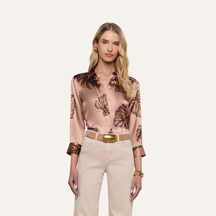 A woman with long blonde hair wears a shiny, light brown Dani Silk Blouse with a dark floral pattern, beige pants, a gold belt, and a gold watch, standing against a plain light background.