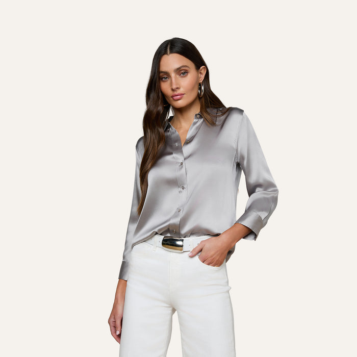 A woman wearing the Dani Silk Blouse, a shiny long-sleeved button-down top, paired with white pants, stands with one hand in her pocket against a plain, light background.
