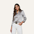 A woman wearing the Dani Silk Blouse, a shiny long-sleeved button-down top, paired with white pants, stands with one hand in her pocket against a plain, light background.