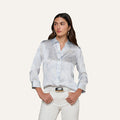 A woman models the Dani Silk Blouse—a light blue patterned silk button-down with rolled sleeves—paired with white pants, standing with her hands in her pockets against a plain light background.