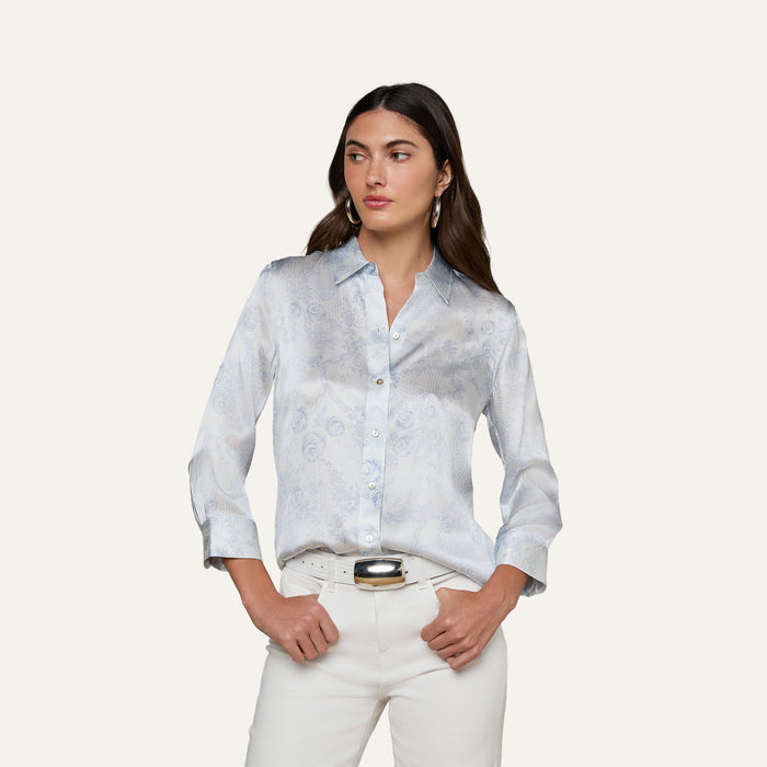 A woman models the Dani Silk Blouse—a light blue patterned silk button-down with rolled sleeves—paired with white pants, standing with her hands in her pockets against a plain light background.
