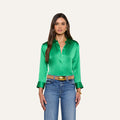 A woman with long brown hair wears the Dani Silk Blouse—shiny green with 3/4 sleeves, wide cuffs, and button-down front—styled with blue jeans, a tan belt featuring a gold buckle, and gold jewelry. She stands against a plain light background.