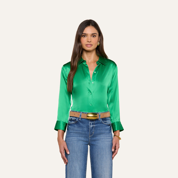 A woman with long brown hair wears the Dani Silk Blouse—shiny green with 3/4 sleeves, wide cuffs, and button-down front—styled with blue jeans, a tan belt featuring a gold buckle, and gold jewelry. She stands against a plain light background.