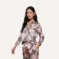 A woman with long dark hair wears a pink, floral-patterned Dani Silk Blouse and matching pants. She stands with one hand in her pocket against a plain, light background.