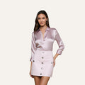 A woman stands against a plain off-white background, wearing a shiny light pink Dani Silk Blouse with rolled sleeves paired with a matching light pink mini skirt adorned with large silver buttons down the front.