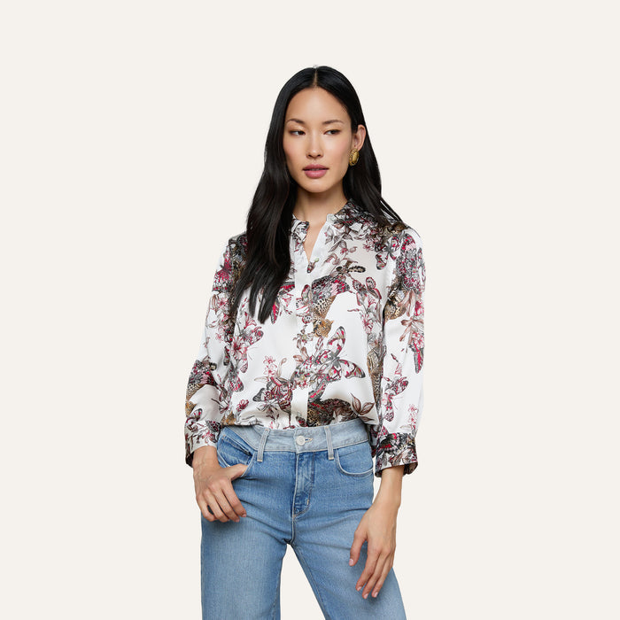 A woman wearing the Dani Silk Blouse—a patterned white top featuring floral, bird, and butterfly jungle print designs—paired with blue jeans stands against a plain background, looking slightly to the side with one hand in her pocket.
