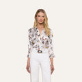 A woman with long blonde hair wears the Dani Silk Blouse, featuring a white silk fabric with a colorful butterfly print, paired with white pants and standing against a plain light background.