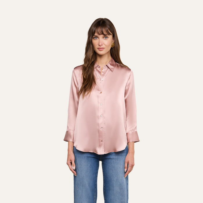 A woman with long brown hair and bangs stands against a plain background, wearing the Dani Silk Blouse—a light pink, 100 silk charmeuse button-up with long sleeves—paired with blue jeans. She has a neutral expression.