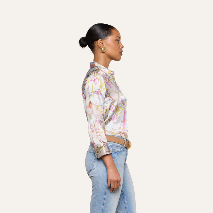 A woman stands in profile against a plain background, wearing a colorful Dani Silk Blouse in a baroque toile print, light blue jeans, gold hoop earrings, and her hair styled in a neat bun.