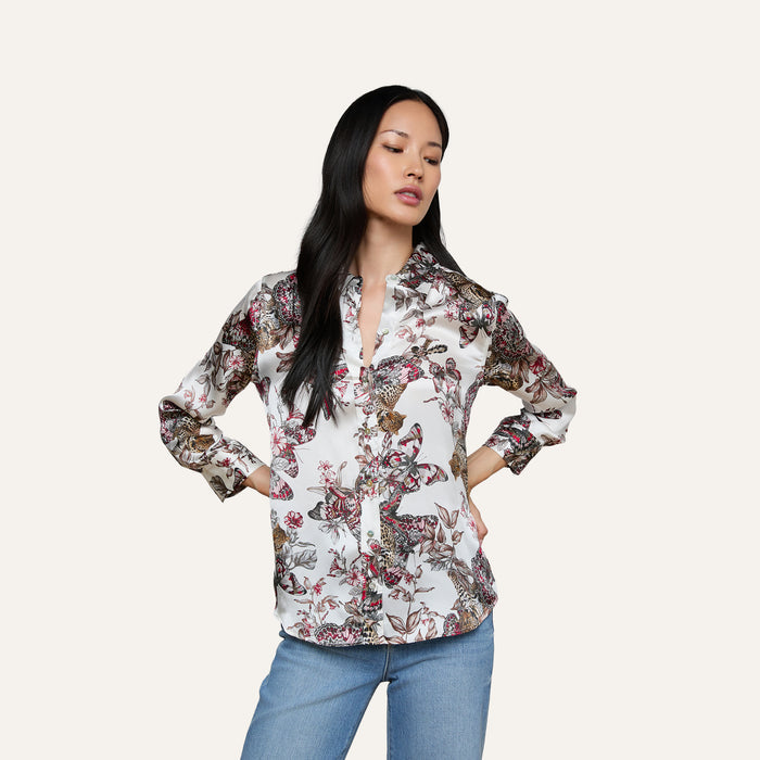A woman with long black hair stands with her hands on her hips against a plain, light background, wearing the Dani Silk Blouse in a butterfly jungle print paired with light blue jeans.