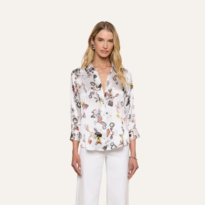 A woman with long blonde hair models the Dani Silk Blouse, featuring a white butterfly print, paired with white pants. She stands against a plain light background and looks at the camera with a slight smile.