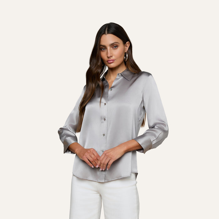 Woman with long brown hair wearing the Dani Silk Blouse and white pants, standing against a plain light background, looking at the camera with hands gently clasped.