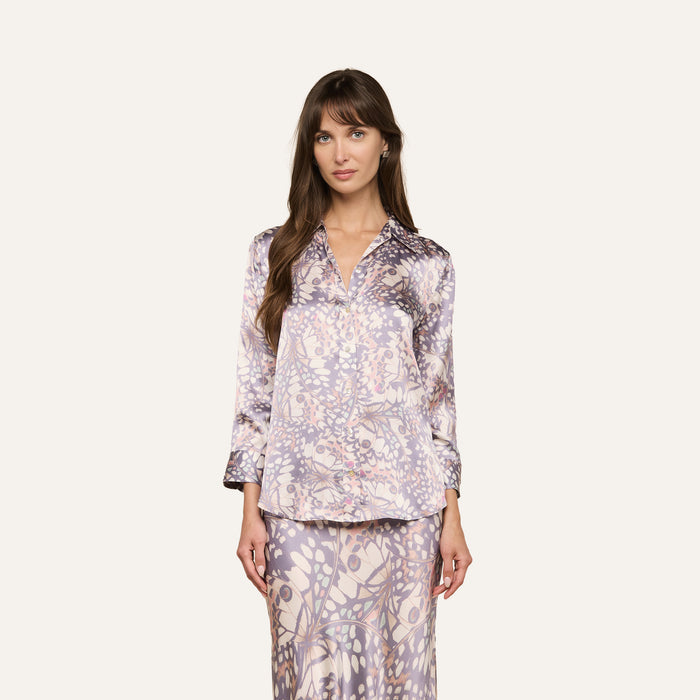 A woman wears the Dani Silk Blouse—a long-sleeve, button-down shirt in silk—paired with a matching skirt. Both pieces showcase a light purple and beige abstract floral pattern reminiscent of butterfly wings, as she stands against an off-white background.