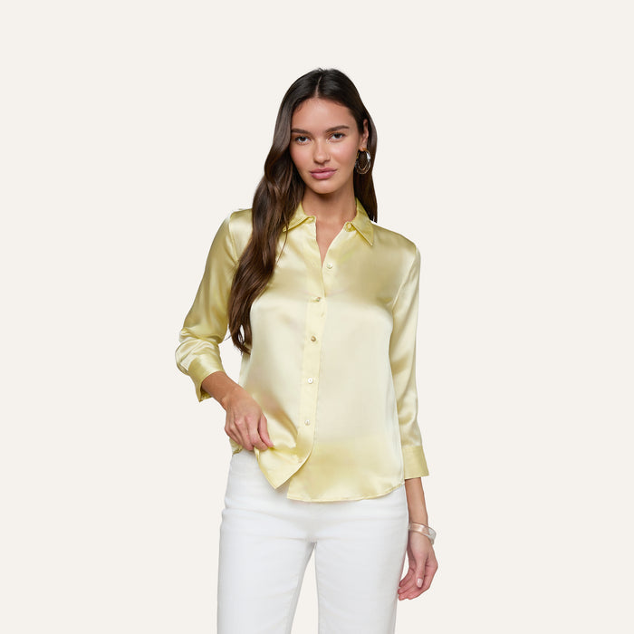 A woman wearing the Dani Silk Blouse in yellow and white pants stands against a plain light background, with one hand in her pocket and the other by her side.