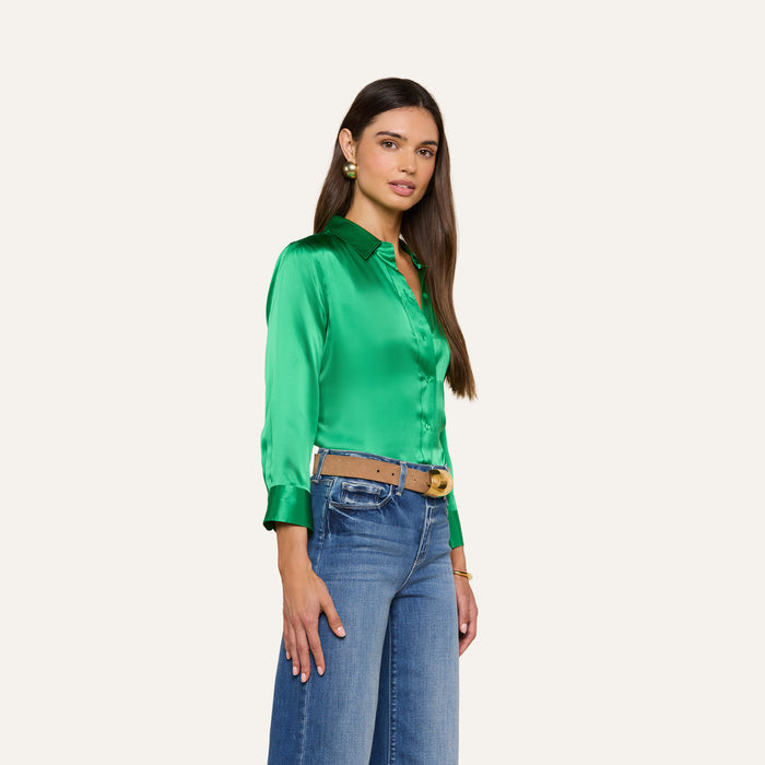 A woman stands facing slightly to the side, wearing a shiny green Dani Silk Blouse with 3/4 sleeves and blue jeans. She has long brown hair, gold earrings, and a tan belt. The background is off-white.
