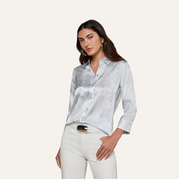 Wearing the Dani Silk Blouse—a light blue patterned silk button-down with 3/4 sleeves—and white pants, a woman stands against a plain off-white background with one hand in her pocket and a neutral expression.