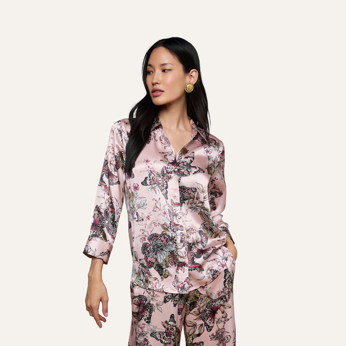 A woman wearing the Dani Silk Blouse in a shiny, light pink silk fabric with a butterfly and floral pattern poses with one hand in her pocket against a plain light background.