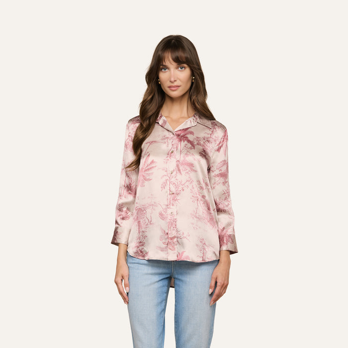 A woman with long brown hair wears a pink floral Dani Silk Blouse with long sleeves and light blue jeans, standing against a plain off-white background.