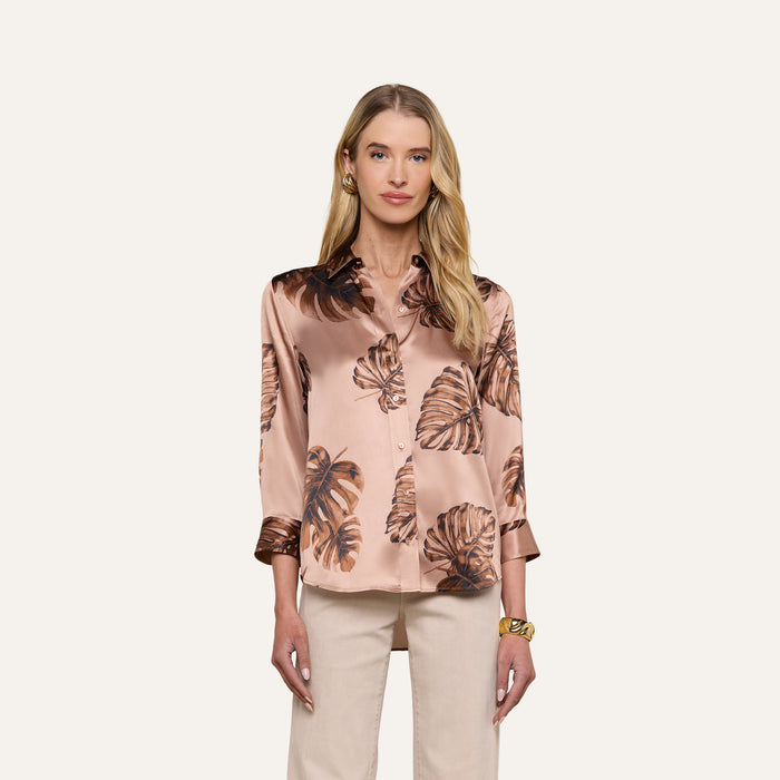 A woman stands facing forward against a plain light background, wearing the Dani Silk Blouse—a light pink, silky top adorned with a brown palm leaf print. She has long blonde hair and pairs the blouse with light-colored pants and a gold bracelet.