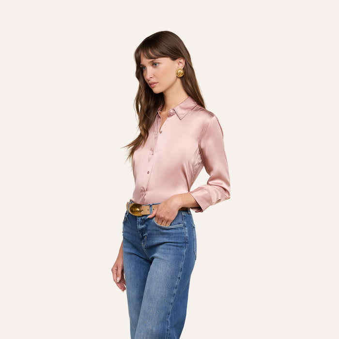 A woman wearing the Dani Silk Blouse in pink, paired with blue jeans and gold earrings, stands against a plain background, looking to the side with one hand on her waist.