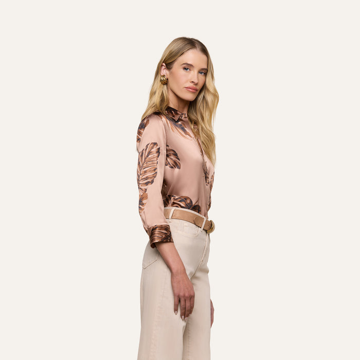 A woman with long blonde hair stands in profile against a plain white background, wearing the Dani Silk Blouse in a pink palm leaf print paired with cream-colored pants.