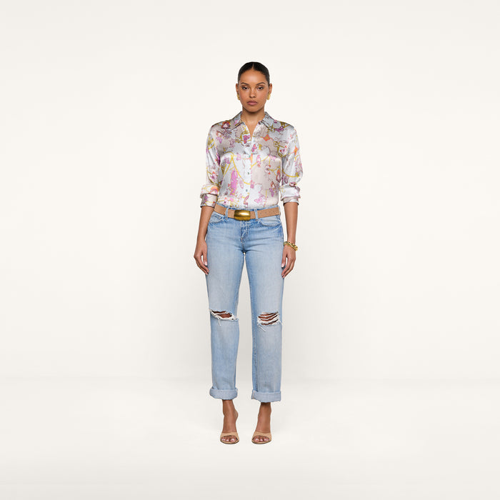 A woman stands against a plain white background, wearing the Dani Silk Blouse in a pastel baroque toile print, paired with light blue ripped jeans, a brown belt, and heeled sandals. Her hair is pulled back and she is facing forward.