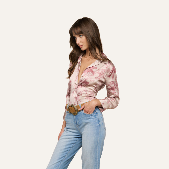 A woman with long brown hair wears the Dani Silk Blouse in a pink jungle toile print paired with light blue jeans, standing with one hand in her pocket and gazing to the side against a plain backdrop.