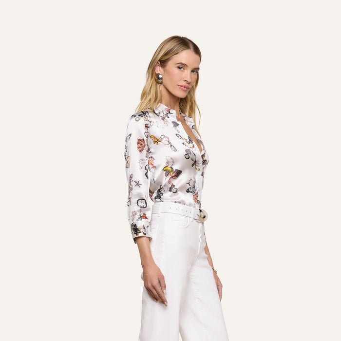 A woman with long blonde hair stands against a plain light background, wearing the Dani Silk Blouse featuring a butterfly print and paired with white pants. She looks slightly to the side with a neutral expression.