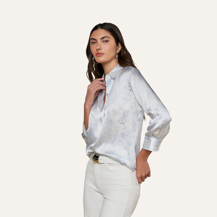 A woman with long brown hair wears the Dani Silk Blouse—a light blue satin button-down featuring subtle floral patterns—paired with white pants against a plain, off-white background.