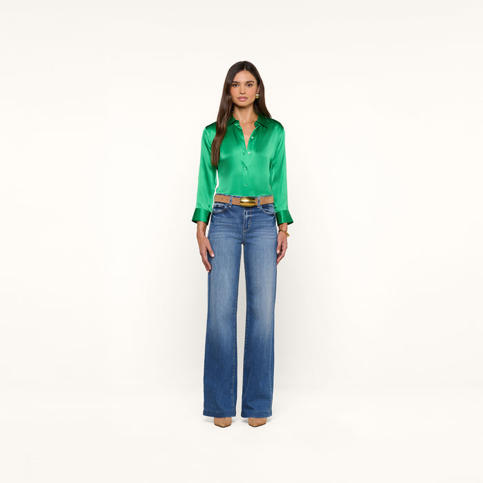 A woman stands facing forward, wearing a shiny green Dani Silk Blouse with 3/4 sleeves, blue high-waisted wide-leg jeans, and tan heels. The background is plain white.