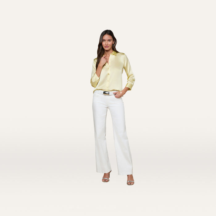 A woman stands against a plain white background, wearing the Dani Silk Blouse in yellow, paired with white pants, heeled sandals, hoop earrings, and a black-buckled belt. She poses with one hand by her chin and the other in her pocket.