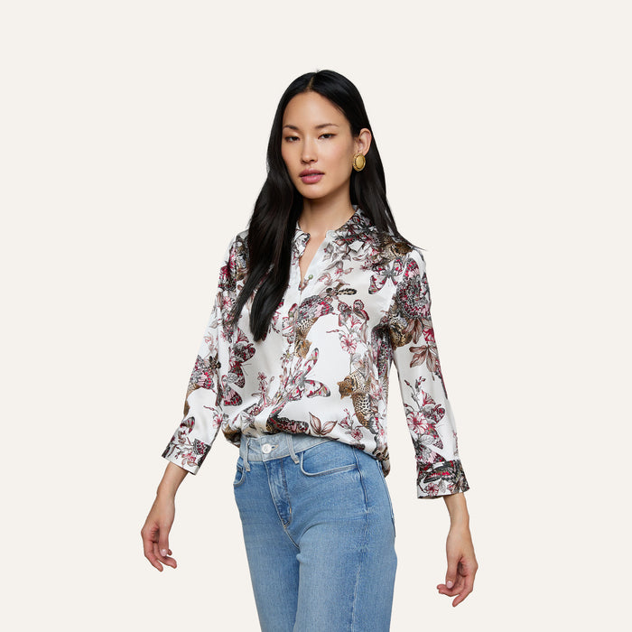 The woman is wearing the Dani Silk Blouse—a white silk button-down featuring a colorful butterfly jungle print—paired with light blue jeans. She has long dark hair and is posing against a plain light background.