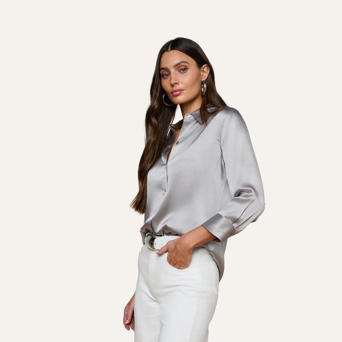 A woman with long brown hair stands confidently against a plain light background, wearing the Dani Silk Blouse in gray paired with white pants, her hand casually in her pocket.