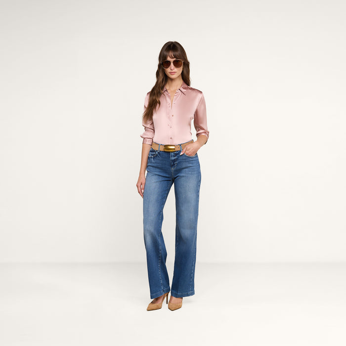 A woman wearing sunglasses, a light pink Dani Silk Blouse, blue high-waisted jeans, a brown belt, and brown heels stands against a plain white background, posing with one hand in her pocket.