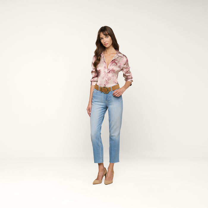 A woman stands in a studio wearing the pink floral Dani Silk Blouse, paired with light blue jeans, a tan belt, and beige heels. She has long wavy hair and looks at the camera with a neutral expression against a white background.