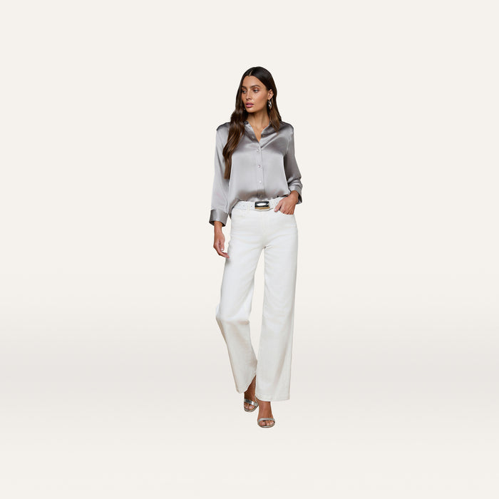 Wearing the Dani Silk Blouse in a shiny silver hue, paired with wide-leg white pants, heels, and drop earrings, a woman stands confidently against a plain light background.