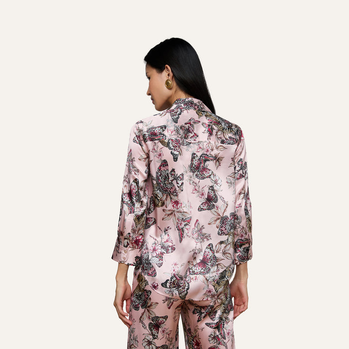 A woman with long dark hair, seen from the back, wears the Dani Silk Blouse featuring a pink butterfly jungle print, standing against a plain light background.