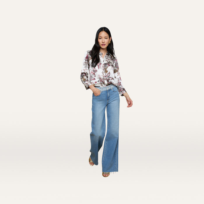 A woman stands confidently against a plain white background, wearing the Dani Silk Blouse featuring a butterfly jungle print and wide-leg blue jeans, with one hand in her pocket and the other relaxed by her side.