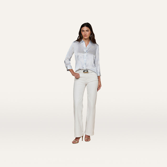 A woman with long dark hair and a neutral expression stands against a plain background wearing the light blue Dani Silk Blouse, paired with white pants, a brown belt, and beige heeled sandals.