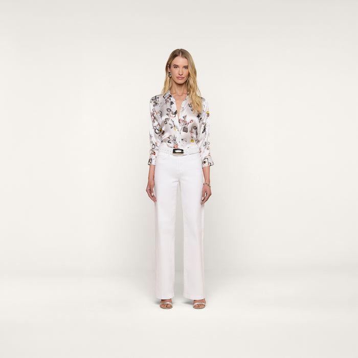 A woman stands facing forward against a plain white background, wearing the Dani Silk Blouse with a butterfly print, tucked into high-waisted white trousers and paired with heeled sandals.