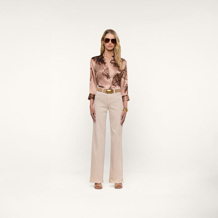 A woman with long blonde hair wears sunglasses, the Dani Silk Blouse featuring a palm leaf print, beige high-waisted pants, and nude heels, standing against a plain white background.