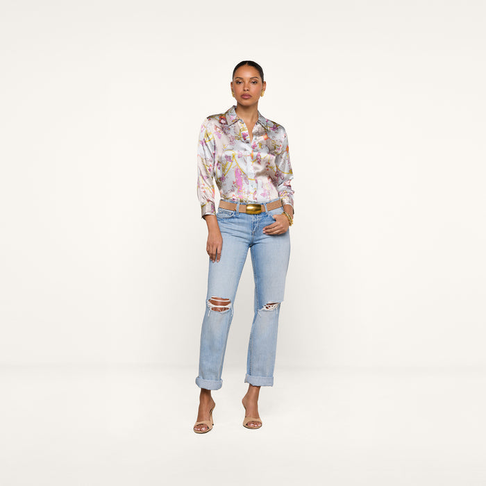 A woman stands against a plain white background, confidently posing with her hands in her pockets. She wears the Dani Silk Blouse in a baroque toile print, styled with light blue ripped jeans, a tan belt, and open-toe heels. Her hair is tied back for a polished yet relaxed look.