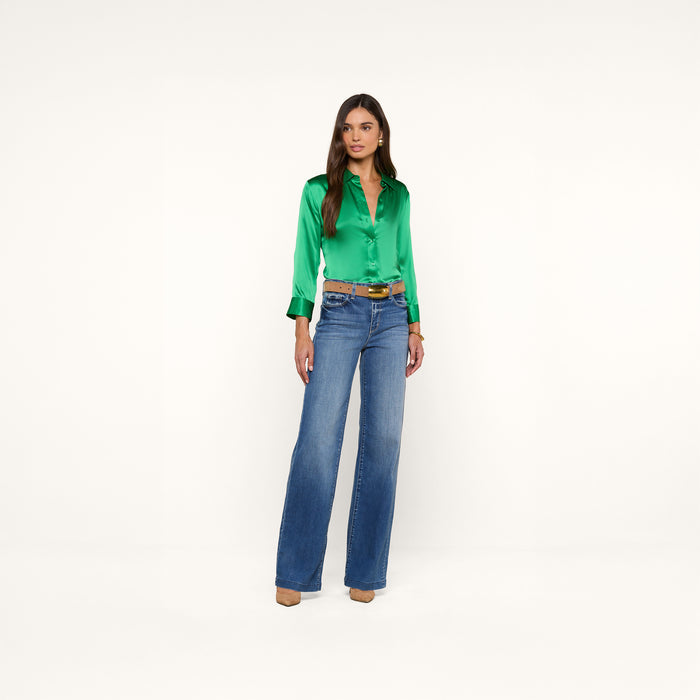 A woman stands against a plain white background, wearing a shiny green Dani Silk Blouse with 3/4 sleeves, blue high-waisted wide-leg jeans, a brown belt, and tan heels. She has long brown hair and a neutral expression.