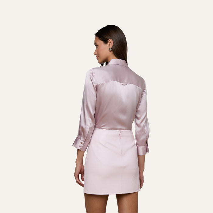 A woman with straight brown hair is shown from behind, wearing a shiny, light pink Dani Silk Blouse with long sleeves and a matching mini skirt. She stands against a plain light background.